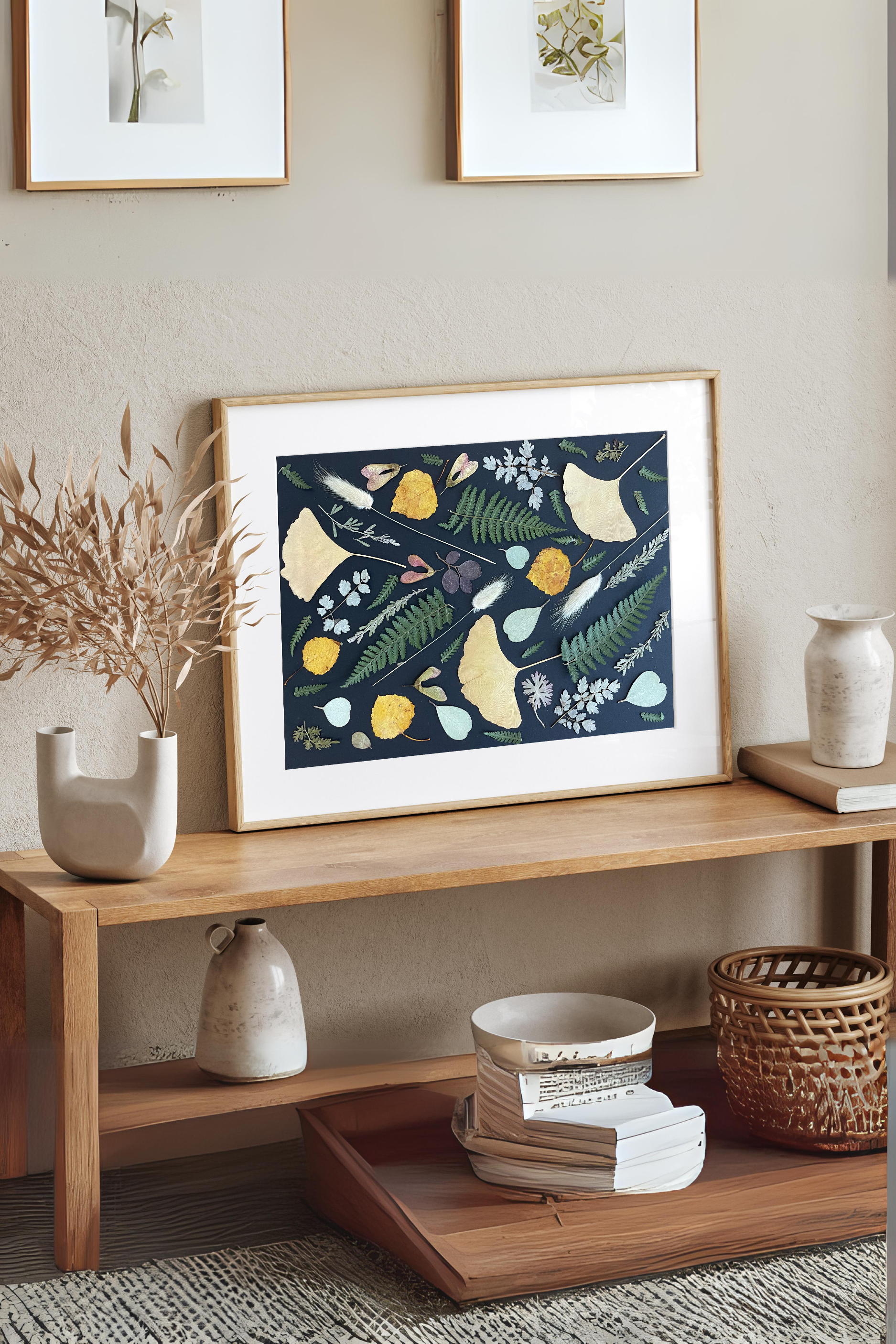 a pressed leaf design with Ginkgo leaves, fern leaves, and fall leaves in a wood frame. Sitting on a soft beige console table.